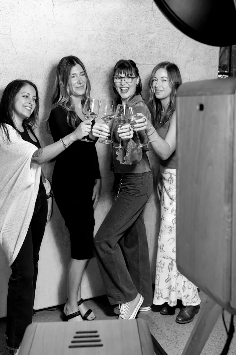 Four women smiling and toasting with drinks in front of a photobooth backdrop.