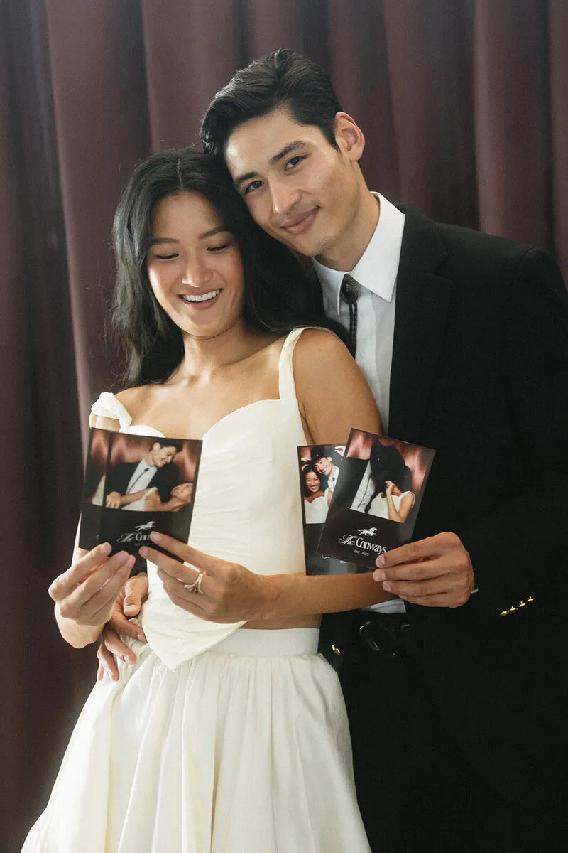 Smiling couple in formal attire holding photobooth prints in front of a dark curtain.