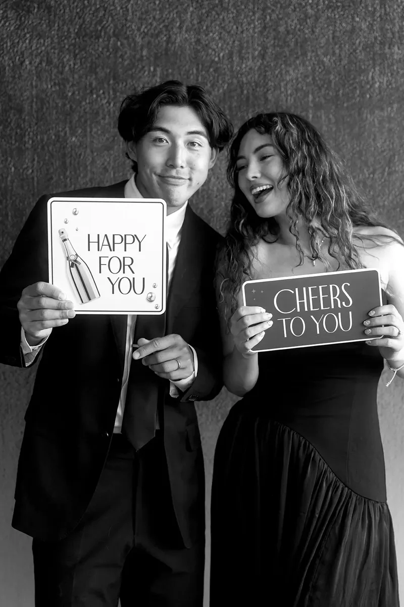 Couple holding photo booth prop signs that read Happy For You and Cheers To You.